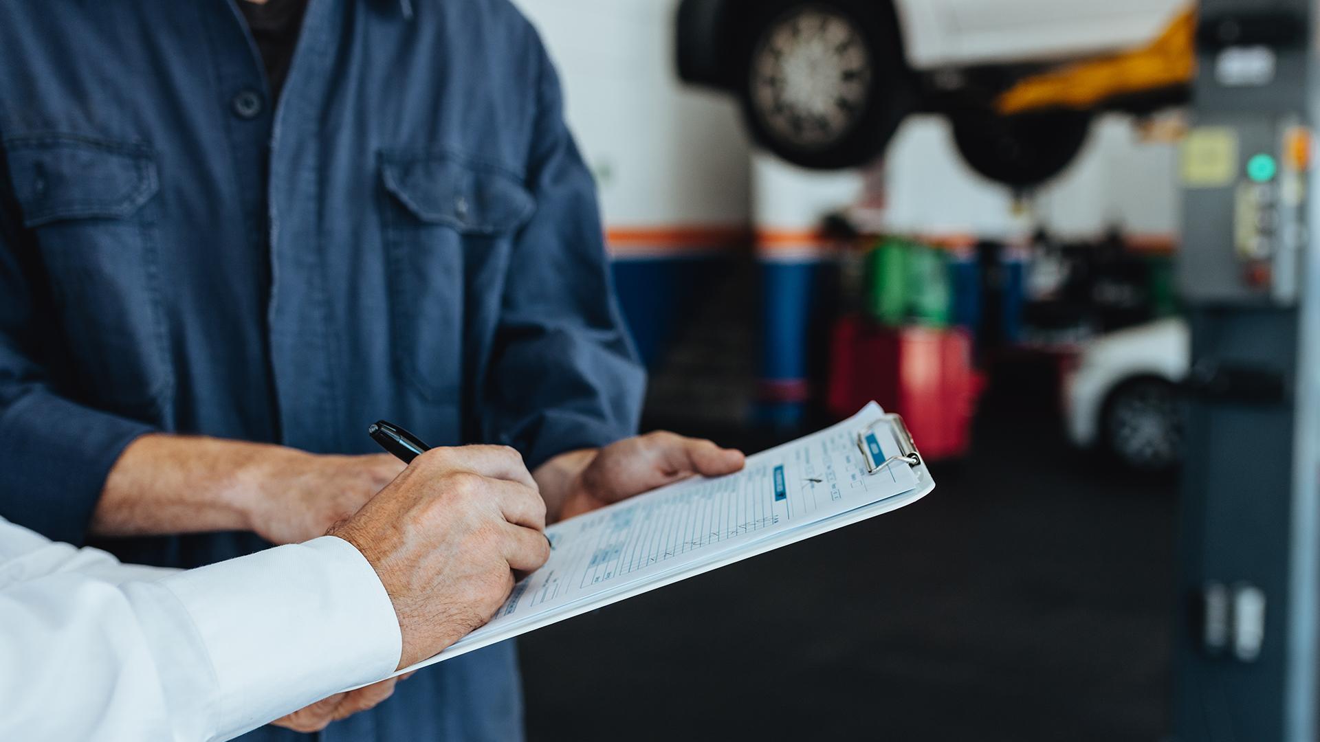 Dans un garage, gros plan sur la main d’un homme qui signe ce qui ressemble à un rapport sur une tablette d’écriture tenue par une personne portant une combinaison de travail.