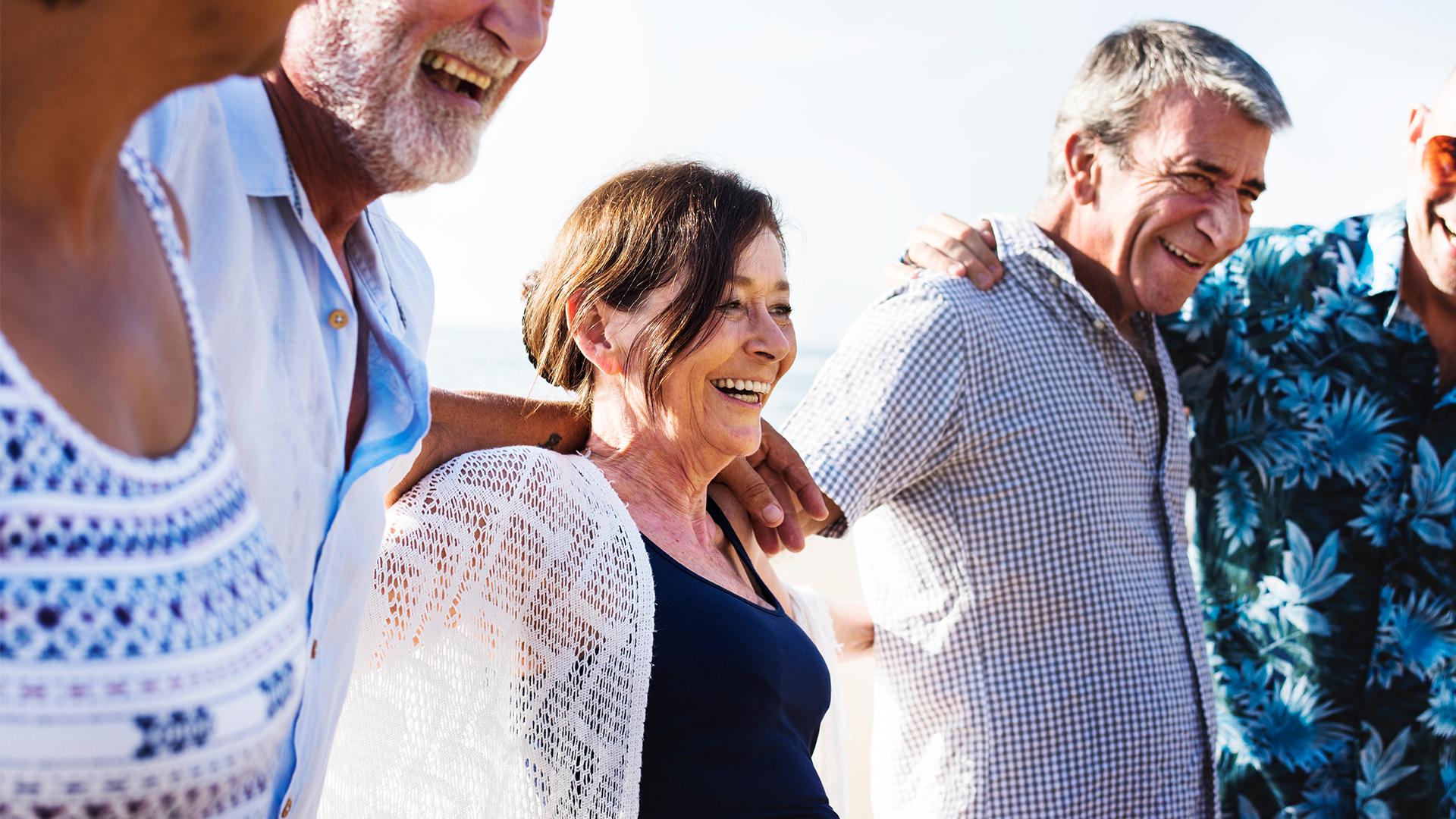 Des snowbirds, hommes et femmes, prennent la pose tout sourire alors qu’ils se promènent au bord de la mer, bras dessus, bras dessous.
