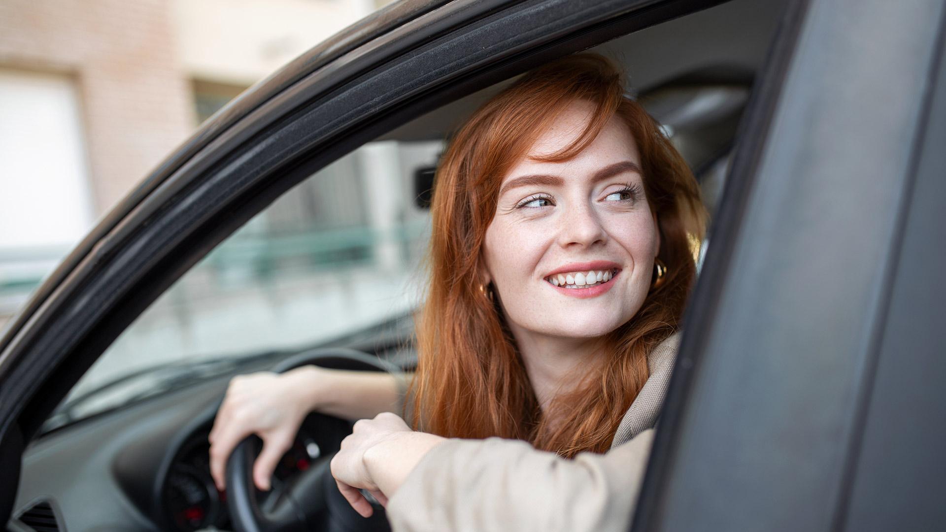 Gros plan sur une femme qui, assise dans sa voiture, regarde par la fenêtre vers l’arrière, une main sur le volant et l’autre appuyée sur le rebord de la fenêtre.