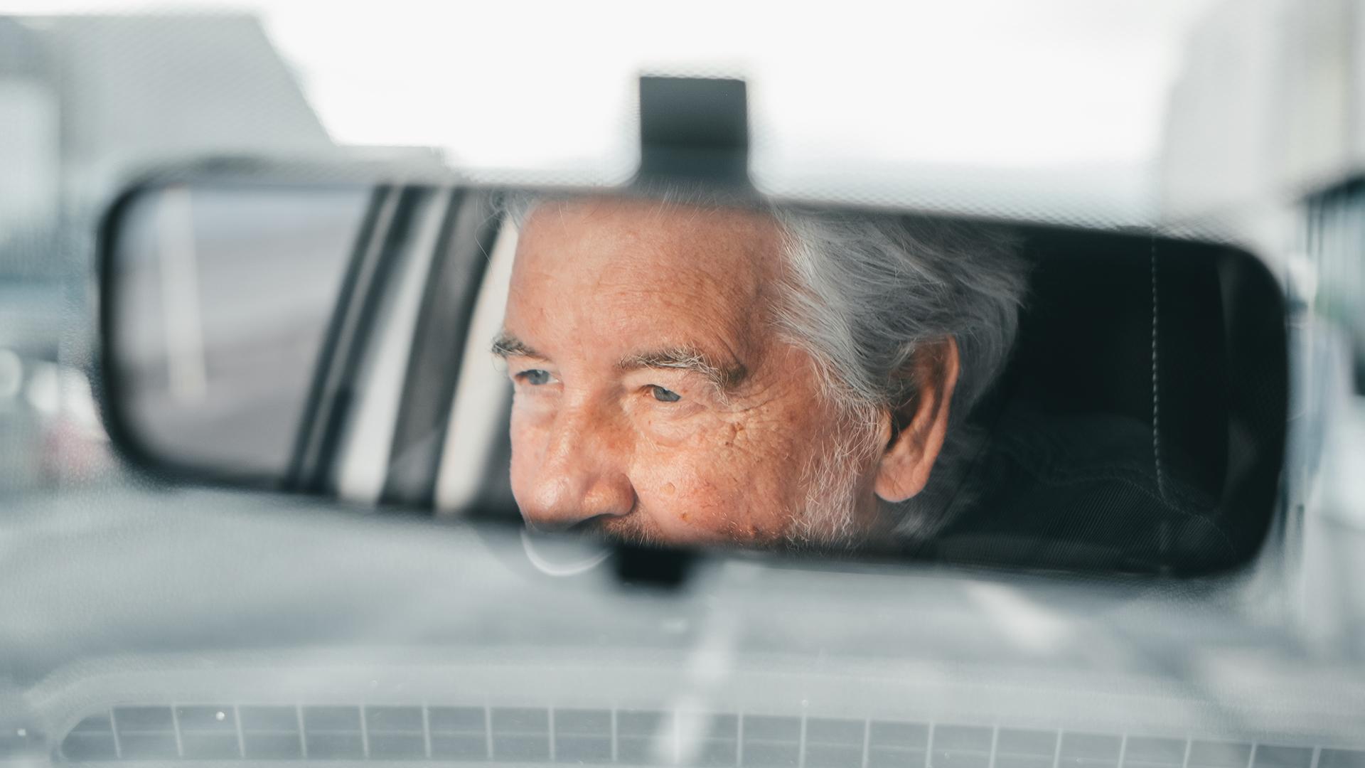 An elderly driver is seen in the central rearview mirror of a car.